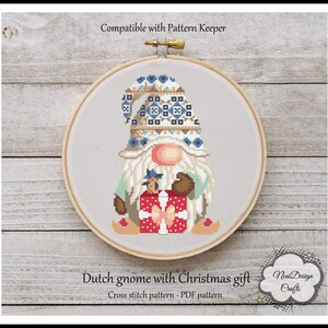 May include: Cross stitch pattern of a Dutch gnome holding a red and white gift box. The gnome is wearing a blue and white hat with a geometric pattern. The pattern is compatible with Pattern Keeper. "Dutch gnome with Christmas gift" and "Cross stitch pattern - PDF pattern" are written below the image.