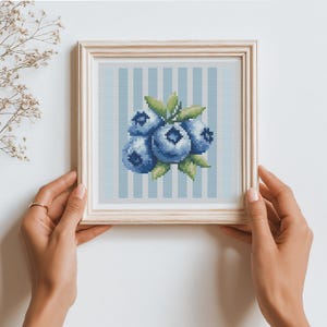 May include: A framed cross-stitch artwork featuring a cluster of blueberries. The blueberries are various shades of blue with green leaves, set against a light blue and white striped background. The frame is a light wood color.