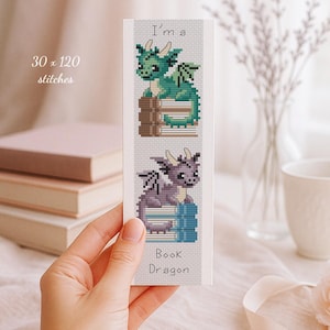 May include: A rectangular cross-stitch pattern bookmark featuring the text I'm a Book Dragon. The design displays a green dragon sitting on brown books and a purple dragon sitting on blue books, with a 30 x 120 stitches measurement label.