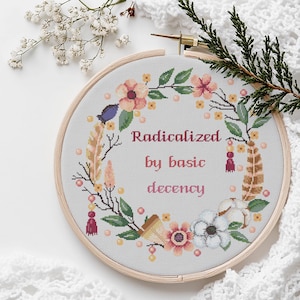 Puede incluir: Un bastidor de bordado de punto de cruz con el texto "Radicalized by basic decency" en un diseño de corona floral. La corona incluye flores rosas y melocotón, hojas verdes y elementos decorativos. El bastidor es de madera.