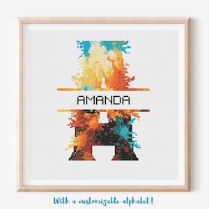 May include: Cross-stitch art featuring the letter "A" in a vibrant, abstract design with orange, yellow, and blue hues. The name "AMANDA" is displayed in black text. The artwork is framed in a light wood frame.