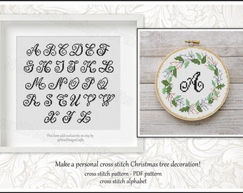 Könnte beinhalten: Ein schwarzes und weißes Kreuzstich-Alphabetmuster mit einer kursivschriftlichen Schriftart. Das Alphabet wird in einem weißen Rahmen angezeigt. Ein Beispiel für ein Kreuzstich-Design wird in einem Reifen mit einem grünen und weißen Kranz und dem Buchstaben "A" in der Mitte gezeigt.