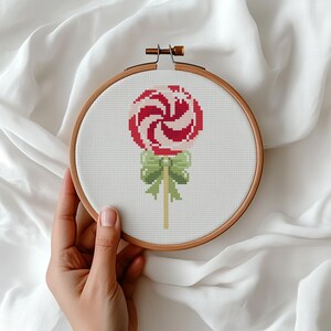May include: A cross-stitch embroidery hoop showing a red and white swirled lollipop design with a green bow and a wooden stick. The embroidery is set against a white fabric background.