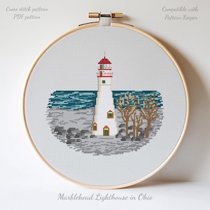 Pode incluir: Padrão de ponto cruz do Farol de Marblehead em Ohio, exibido em um bastidor de bordado de madeira. O farol é branco com um topo vermelho, contra um mar azul e rochas cinzentas. O texto "Cross stitch pattern PDF pattern" e "Compatible with Pattern Keeper" são visíveis.