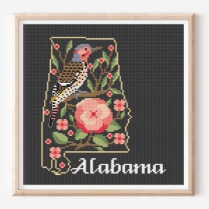 May include: A framed cross-stitch embroidery featuring the outline of the state of Alabama on a dark gray background. Inside the outline is a northern flicker bird perched on a branch with pink camellia flowers and the word Alabama in white.