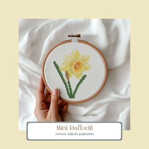 May include: A cross-stitch pattern of a yellow daffodil with green leaves and stem, displayed in a wooden embroidery hoop. The text "Mini Daffodil" and "cross stitch pattern" are below the hoop.