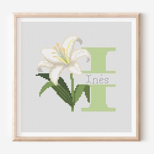 May include: Cross-stitch artwork in a wooden frame. The design shows a white lily with green leaves and a light green letter "I". The name "Inés" is stitched below the letter. The background is a light grey.
