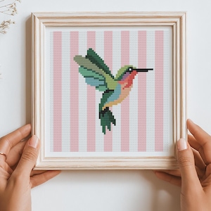 Pode incluir: Um bordado em ponto cruz quadrado apresentando um beija-flor pixelizado com plumagem verde, azul e rosa, centralizado contra um fundo de listras verticais rosa claro e branco, inserido em uma moldura de madeira de cor clara.