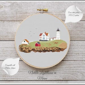 May include: Cross-stitch pattern of the Nubble Lighthouse in Maine, featuring a white lighthouse, a red-roofed house, and a small red building on a green hill. The design is set against a light gray background within a wooden embroidery hoop.