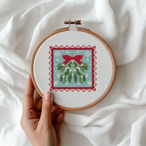 May include: A cross-stitch embroidery hoop featuring a festive mistletoe design. The design includes a red bow, green mistletoe leaves, and white snowflakes on a light blue background, framed by a red border. The wooden hoop is held by a hand.