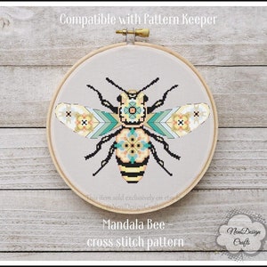 Puede incluir: Un patrón de punto de cruz de una abeja con un diseño de mandala. La abeja es amarilla y negra con alas azul verdosas y blancas. El patrón es compatible con Pattern Keeper. "Mandala Bee - cross stitch pattern" está escrito debajo de la abeja.