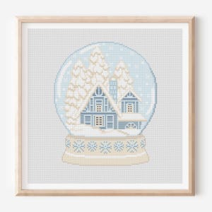May include: A framed cross-stitch of a snow globe featuring a blue house and snow-covered trees. The snow globe is set against a light blue background with a beige base and a wooden frame. The design has a winter theme.