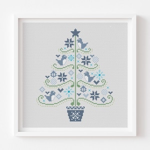 May include: A framed cross-stitch art piece featuring a stylized Christmas tree design. The tree is composed of blue, green, and white cross-stitch elements, including birds, snowflakes, and geometric shapes. The artwork is set against a light gray background.