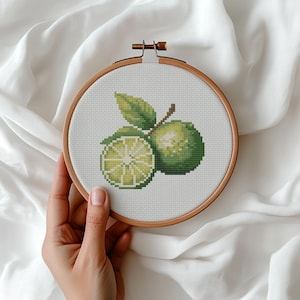 May include: A cross-stitch embroidery hoop featuring a green lime design. The design includes a whole lime and a lime cut in half, with green leaves. The embroidery is set in a wooden hoop, showcasing a handmade craft project.