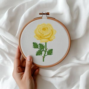 May include: A cross-stitch embroidery hoop featuring a yellow rose. The rose is in full bloom with green leaves and stem. The embroidery is set against a white background, framed by a wooden hoop.