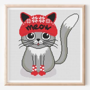 Könnte beinhalten: Ein gerahmtes Kreuzstichbild einer grauen Katze mit roter Mütze mit dem Wort "meow" und Schneeflocken. Die Katze trägt auch rote Socken mit weißen Mustern. Das Kunstwerk hat einen hellgrauen Hintergrund und einen Holzrahmen.