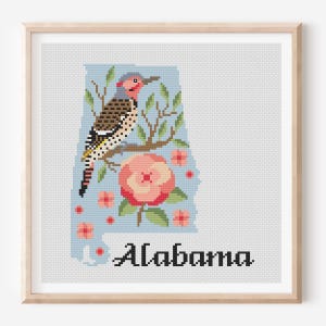 Könnte beinhalten: Eine gerahmte Kreuzstichstickerei, die den Bundesstaat Alabama in Hellblau darstellt, verziert mit einem Goldspecht auf einem braunen Ast, rosa Kamelienblüten mit grünen Blättern und dem Wort Alabama in Schwarz, in einem hellen Holzrahmen.