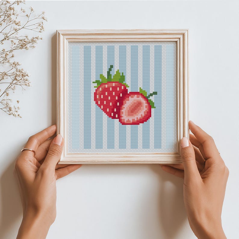 May include: A framed cross-stitch artwork featuring two red strawberries with green leaves against a light blue and white striped background. The artwork is in a light wooden frame.