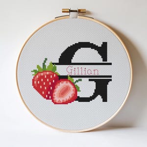 Op de afbeelding: Kruissteekborduurwerk in een houten hoepel. Het ontwerp toont een grote zwarte letter "G" met de naam "Gillian" in roze. Twee rode aardbeien met groene bladeren staan links van de letter.