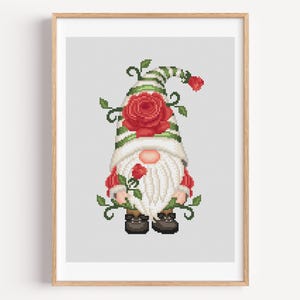 May include: Cross-stitch art of a garden gnome with a white beard, a red nose, and a striped green and white hat. The gnome is holding a red rose and surrounded by green vines and red roses. The artwork is framed in a light brown wooden frame.