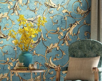 May include: A teal blue armchair with a beige pillow sits in front of a wall with a blue and gold wallpaper featuring a baroque pattern. A small gold metal side table with two shelves holds a vase with yellow flowers and a stack of books.