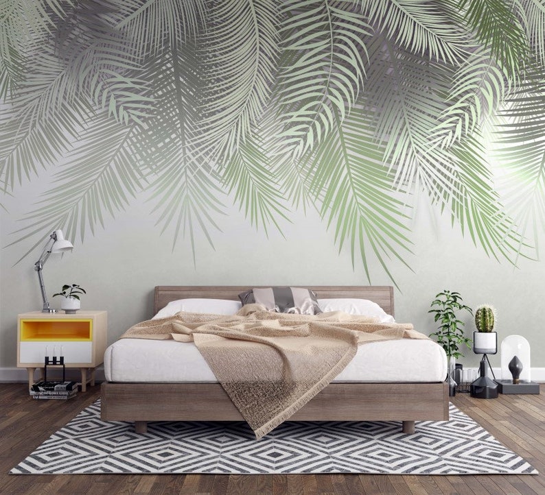 K&ouml;nnte beinhalten: Ein Schlafzimmer mit einem Bett, einem Nachttisch und einem Teppich. Die Wand ist mit einer gro&szlig;fl&auml;chigen Tapetenmusterung aus Palmbl&auml;ttern verziert. Das Bett hat eine wei&szlig;e Bettdecke und eine braune Tagesdecke. Der Nachttisch ist gelb und wei&szlig; und hat eine Lampe darauf. Der Teppich ist schwarz und wei&szlig; und hat ein geometrisches Muster.