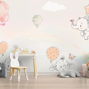 May include: A nursery room with a white wall decorated with colorful hot air balloons and elephants. The wall has a rainbow effect. There is a wooden chair with a bunny rabbit design, a panda-shaped storage bin, and a wooden table with a lamp.