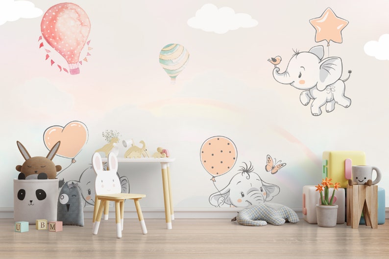May include: A nursery room with a white wall decorated with colorful hot air balloons and elephants. The wall has a rainbow effect. There is a wooden chair with a bunny rabbit design, a panda-shaped storage bin, and a wooden table with a lamp.