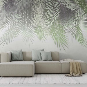 K&ouml;nnte beinhalten: Ein hellgraues Ecksofa mit drei Kissen und einer Wolldecke auf der Chaiselongue. Das Sofa steht vor einer Wand mit einem gro&szlig;fl&auml;chigen Druck von Palmbl&auml;ttern in Gr&uuml;n- und Graut&ouml;nen.