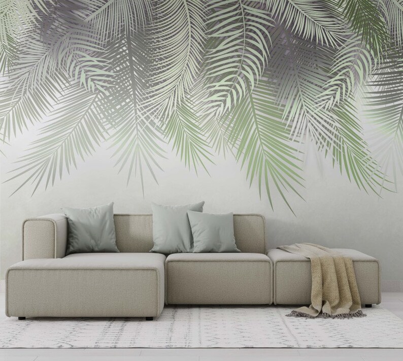 K&ouml;nnte beinhalten: Ein hellgraues Ecksofa mit drei Kissen und einer Wolldecke auf der Chaiselongue. Das Sofa steht vor einer Wand mit einem gro&szlig;fl&auml;chigen Druck von Palmbl&auml;ttern in Gr&uuml;n- und Graut&ouml;nen.