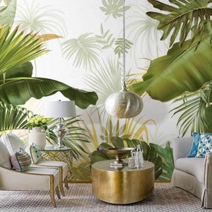 May include: A living room interior design with a white wall featuring a tropical leaf wallpaper. The room has two white armchairs, a gold round coffee table, a gold lamp with a white shade, and a silver pendant light.