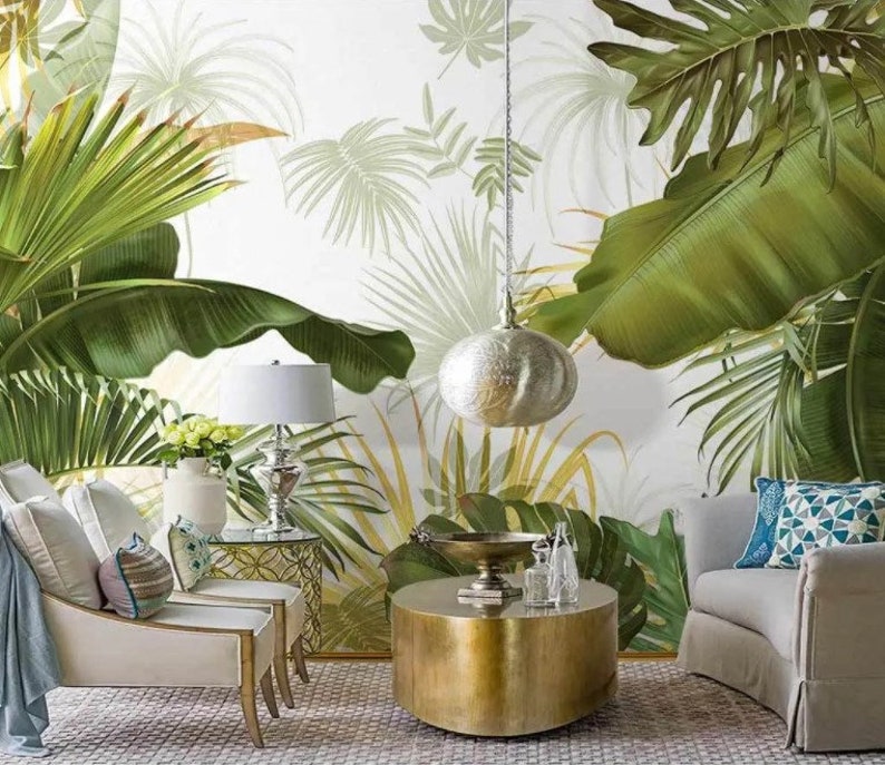 May include: A living room interior design with a white wall featuring a tropical leaf wallpaper. The room has two white armchairs, a gold round coffee table, a gold lamp with a white shade, and a silver pendant light.