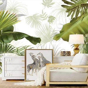 May include: A white dresser with three drawers sits in front of a white wall with a tropical leaf pattern. A white armchair with a gold frame sits in front of the dresser. A framed print of giraffes hangs above the dresser. A lamp with a white shade sits on a table next to the armchair.