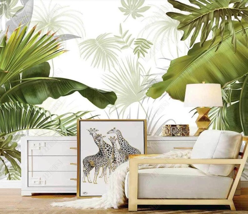 May include: A white dresser with three drawers sits in front of a white wall with a tropical leaf pattern. A white armchair with a gold frame sits in front of the dresser. A framed print of giraffes hangs above the dresser. A lamp with a white shade sits on a table next to the armchair.