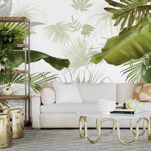 May include: A white sofa with two white pillows and a brown and beige pillow sits in front of a wall with a tropical leaf wallpaper. A gold metal coffee table with a white marble top sits in front of the sofa. Two gold metal stools are in the foreground.