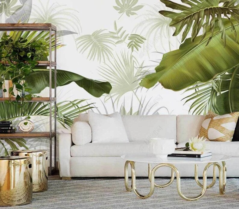 May include: A white sofa with two white pillows and a brown and beige pillow sits in front of a wall with a tropical leaf wallpaper. A gold metal coffee table with a white marble top sits in front of the sofa. Two gold metal stools are in the foreground.