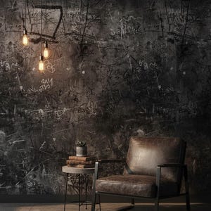 Puede incluir: Escena de diseño de interiores con una pared oscura y texturizada con marcas tipo graffiti. Un sillón de cuero marrón con estructura de metal negro sobre un suelo embaldosado. Una pequeña mesa auxiliar con libros y una planta en maceta, con bombillas colgantes.