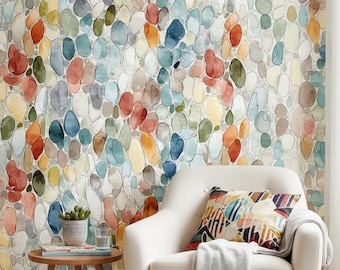 May include: A cream-colored armchair with a patterned throw pillow and a fringed blanket, positioned in front of wallpaper featuring a repeating watercolor pebble design in shades of blue, orange, green, and gray, next to a small wooden side table.
