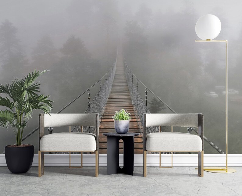 May include: Two white chairs with gold legs sit in front of a foggy forest scene with a suspension bridge leading into the distance. A floor lamp with a white globe sits to the right of the chairs.