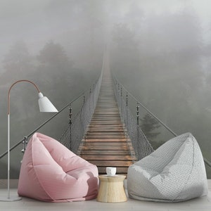 May include: A foggy forest scene with a suspension bridge leading into the distance. Two bean bag chairs are placed in front of the image, one pink and one gray, with a small wooden table and a white cup in between.
