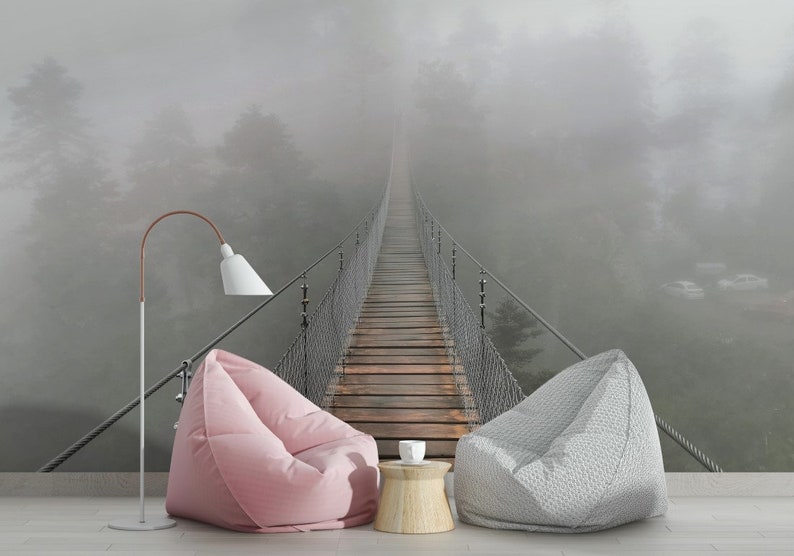 May include: A foggy forest scene with a suspension bridge leading into the distance. Two bean bag chairs are placed in front of the image, one pink and one gray, with a small wooden table and a white cup in between.