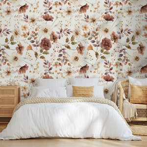 Puede incluir: Una cama blanca con una funda n&oacute;rdica blanca y una manta de punto beige. La cama est&aacute; frente a una pared con un papel tapiz floral con un patr&oacute;n de flores marrones, blancas y moradas. Una silla de mimbre beige con un coj&iacute;n beige est&aacute; a la derecha de la cama.