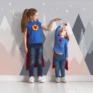 May include: Two children dressed as superheroes stand in front of a wall with a mountain and star pattern. The taller child is wearing a blue shirt with a red star and a red cape. The shorter child is wearing a blue shirt with a red star and a red cape.