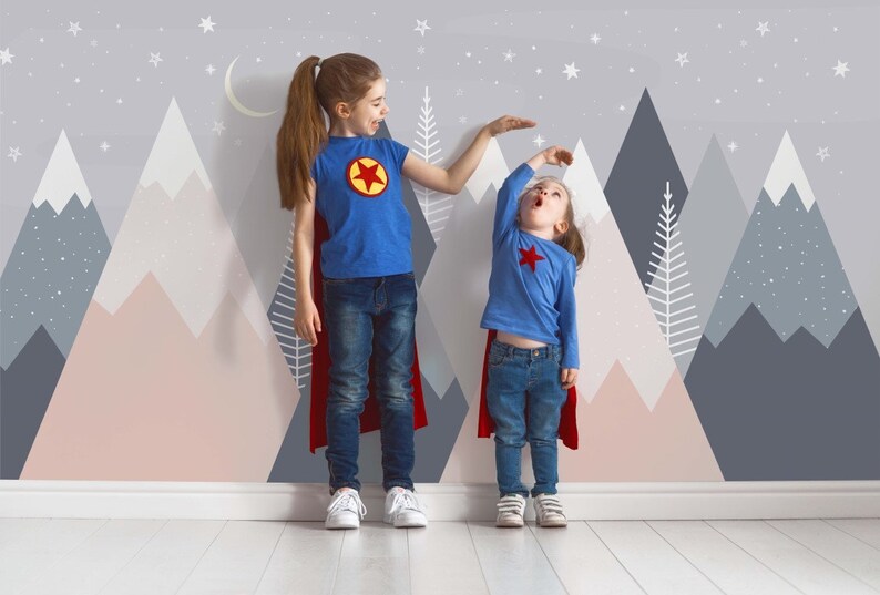 May include: Two children dressed as superheroes stand in front of a wall with a mountain and star pattern. The taller child is wearing a blue shirt with a red star and a red cape. The shorter child is wearing a blue shirt with a red star and a red cape.