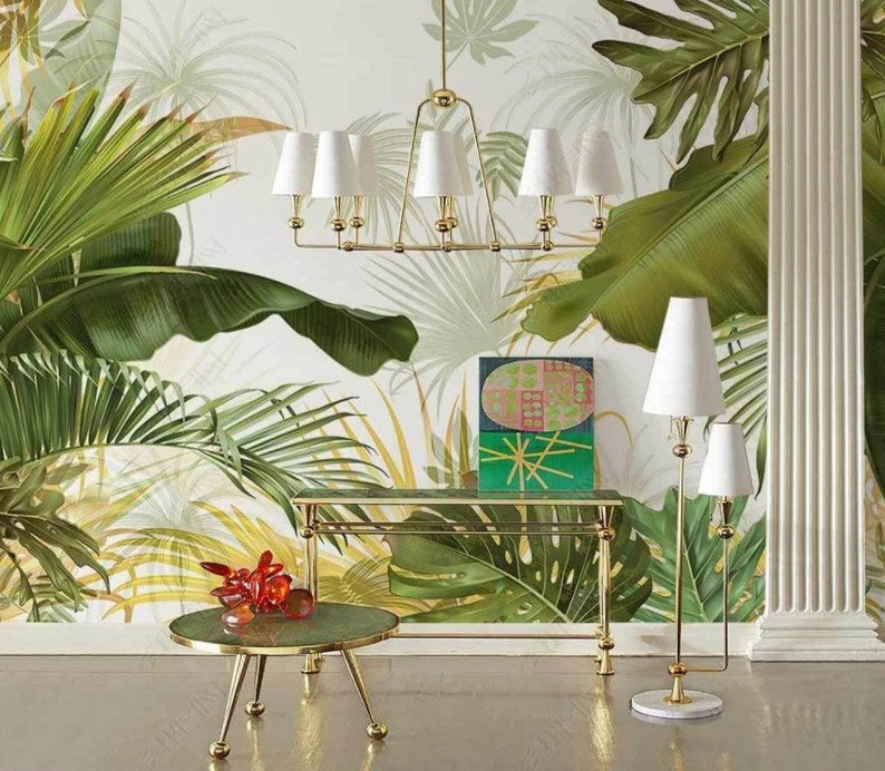 May include: A gold metal table lamp with a white shade and a white marble base. The lamp is next to a gold metal table with a green glass top. The table is decorated with a red glass sculpture. The background is a white wall with a tropical leaf pattern.