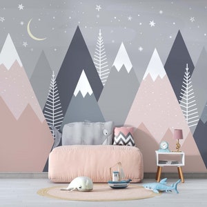 May include: A pink and gray mountain-themed nursery wall mural with a crescent moon and stars. A pink bed with a gray and white pillow and a gray and white chevron pillow is in front of the mural. A small white side table with a lamp and a clock is next to the bed. A blue toy shark and a white toy seal are on the floor in front of the bed.