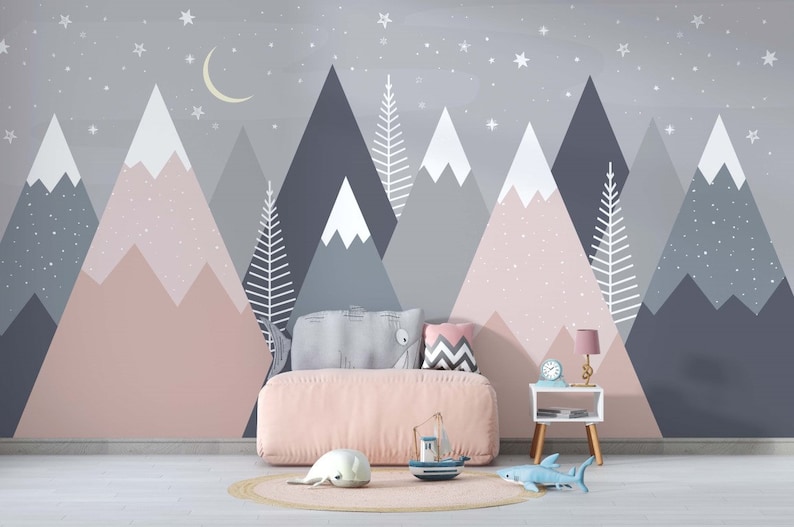 May include: A pink and gray mountain-themed nursery wall mural with a crescent moon and stars. A pink bed with a gray and white pillow and a gray and white chevron pillow is in front of the mural. A small white side table with a lamp and a clock is next to the bed. A blue toy shark and a white toy seal are on the floor in front of the bed.