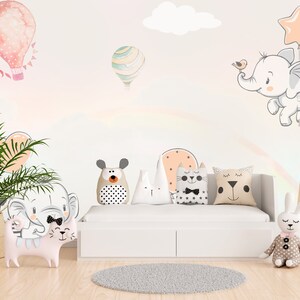 May include: A white couch with a rainbow design on the wall behind it. There are several stuffed animals on the couch, including a cat, a bear, a rabbit, and an elephant. There is also a plant in a woven basket in the corner of the room.