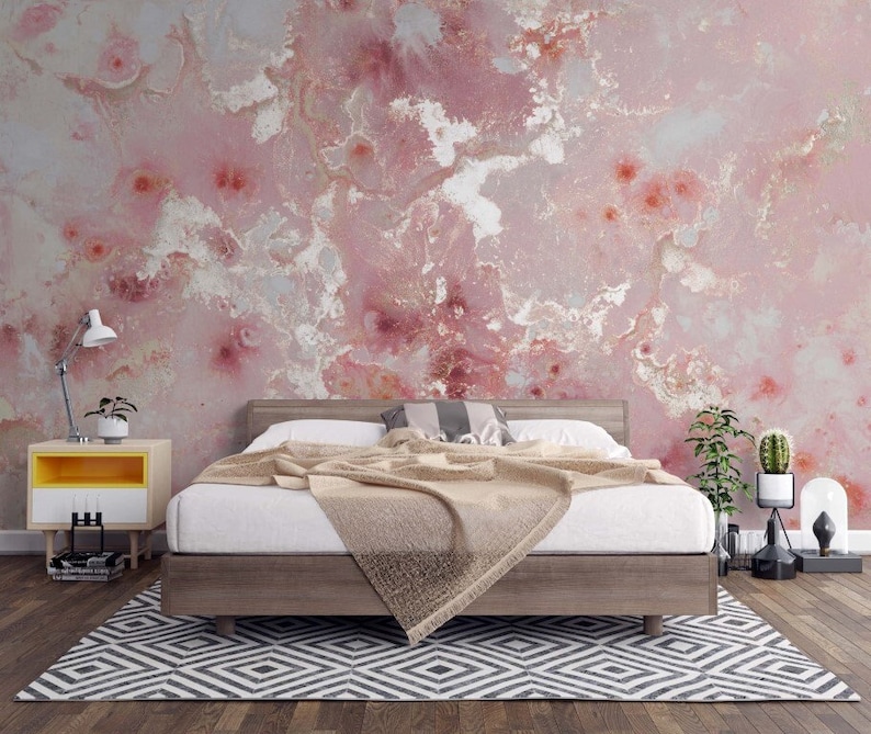 May include: A bedroom with a pink and white marble patterned wallpaper, a wooden bed frame with a white duvet and a beige throw blanket, a wooden nightstand with a yellow drawer, a lamp, and a potted plant.