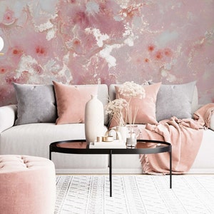 May include: A white sofa with pink and gray throw pillows sits in front of a pink and white abstract wall. A pink throw blanket is draped over the sofa. A black coffee table with a glass top sits in front of the sofa. A pink ottoman sits in front of the coffee table. A white rug with a black and white pattern is on the floor.